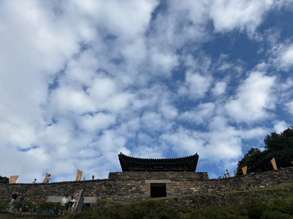 Looking up from below Gongsanseong Fortress in Gongju, with a stone wall and traditional gate pavilion silhouetted against a wide blue sky filled with fluffy clouds