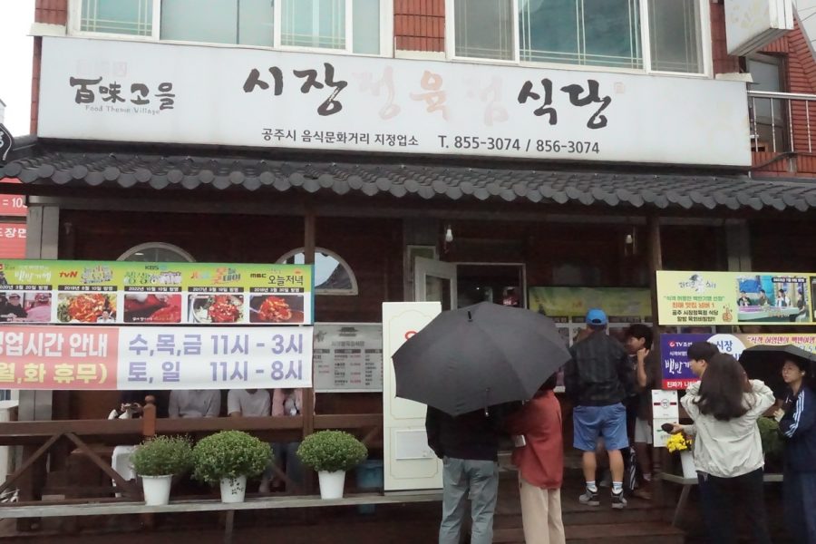 Exterior of Sijang Jeongyukjeom Sikdang in Gongju, with signs in Korean and customers waiting outside under umbrellas.