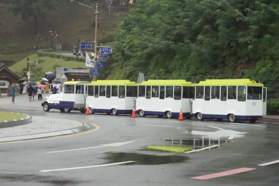 The Goma Train tourist trolley in Gongju parked on a rainy road near major attractions, with visitors walking nearby under umbrellas.